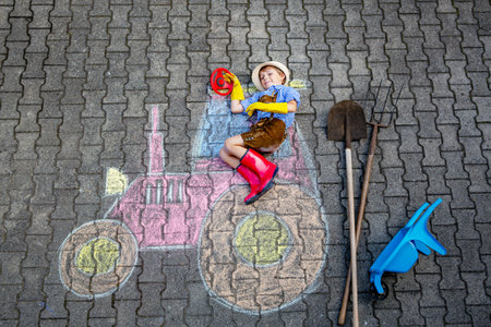 Happy little kid boy in straw hat and rain boots having fun with tractor picture drawing with colorful chalks. Children, lifestyle, fun concept. child dreaming of future and professionの写真素材
