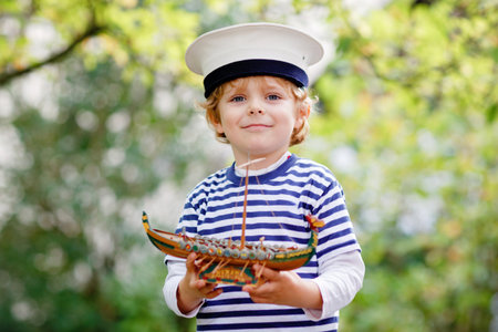 Happy little kid boy in sailor capitain hat and uniform playing with sailor boat ship. Smiling preschool child dreaming and having fun. Education, profession, dream conceptの写真素材