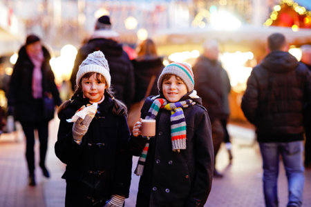 Cute little kids girl and boy having fun on traditional Christmas market during strong snowfall. Happy children eating traditional curry sausage called wurst and drinking hot chocolate. Twins friendsの写真素材