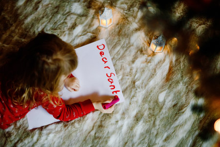 Little cute preschool girl in pajamas under Christmas tree, writing wish list letter to Santa Claus at home, indoors. Traditional Christian festival. Happy toddler child waiting for gifts on xmas.の写真素材
