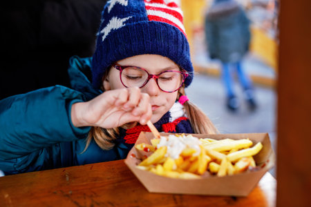 Portrait of happy smiling preschool girl eating french fries with tomato ketchup on Christmas market in Germany outdoors. Little child with eyeglasses enjoy unhealthy fast food or fresh prepared lunchの写真素材