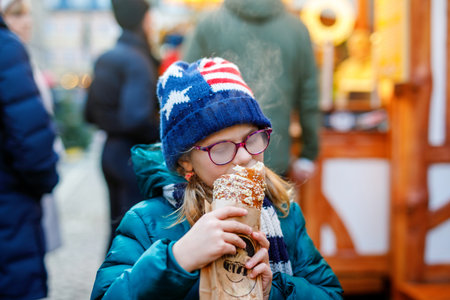 Little school girl, cute child eating sweet bread bun with decoration and lights on background. Happy child on Christmas market in Germany.の写真素材