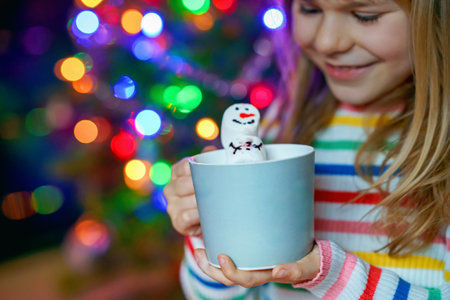 Little child girl holding cup with hot chocolate with marshmallows as snowman. Kid sitting near Christmas tree decorated with lights.の写真素材