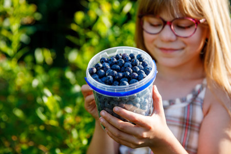 Little schoolgirl with glasses picking and eating fresh ripe blueberries in summer. Happy child enjoying nature. Real life, real people.の写真素材