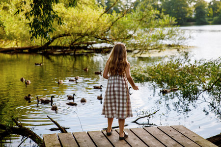 Little elementary school girl feeding wild ducks on a pond. Cute child enjoying nature and animals outdoors in summer, happy kid with eye glasses spending time near water.の写真素材