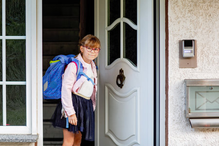 Happy little elementary school girl with eyeglasses and satchel backpack leaving home on the way to school. Smiling pupil child ready for back to school, education and childhood lifestyle concept.の写真素材