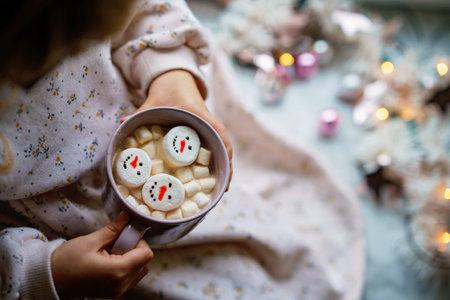 Unrecognizable little child girl holding cup with hot chocolate with marshmallows as snowman. Kid...