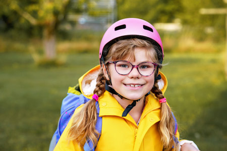 Happy elementary school girl riding a scooter to school wearing a helmet and backpack. Safe, active, real-life cute child commuting in the city.の写真素材