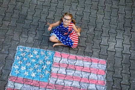 Little girl with glasses and stars and stripes dress, sits on a paved surface beside a large chalk...