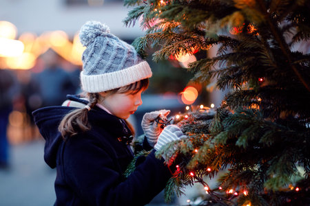 Little cute kid girl having fun on traditional Christmas market during strong snowfall. Happy child enjoying traditional family market in Germany. Schoolgirl standing by illuminated xmas treeの写真素材