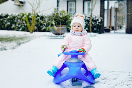 Cute little toddler girl enjoying a sleigh ride on snow. Child sledding. Baby kid riding a sledge in colorful fashion clothes. Outdoor active fun for family winter vacation on day with snowfall.の写真素材