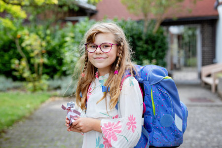 Cute little elementary school girl with eyeglasses and satchel backpack walking outdoors on the way to school. Smiling pupil child ready for back to school, education and childhood lifestyle concept.の写真素材