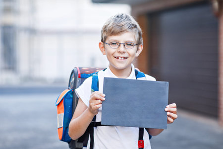 Happy little kid boy with eyeglasses and satchel. Schoolkid on the way to school. Healthy child outdoors holding chalk desk for copyspace. Back to school or school's out.の写真素材