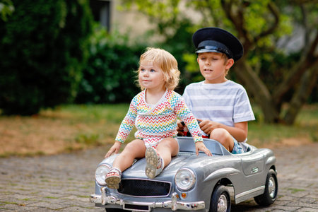 Two happy children playing with big old toy car in summer garden, outdoors. Kid boy driving car with little toddler girl inside. Laughing and smiling kids. Family, childhood, lifestyle concept.の写真素材