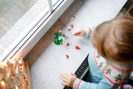 Little toddler girl sitting by window and decorating small glass Christmas tree with tiny xmas toys. Happy healthy child celebrate family traditional holiday. Adorable babyの写真素材
