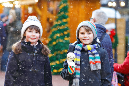Little cute kid girl and boy eating white chocolate covered strawberries and apple on skewer at traditional German Christmas market. Happy children, best friends, twins and siblings on snowy day.の写真素材