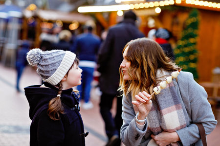 Young mother and daughter eating white chocolate covered fruits and strawberry on skewer at traditional German Christmas market. Happy girl and woman on traditional family market in Germany during snowy dayの写真素材