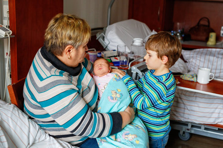 Happy grandmother holding newborn baby in hospital with proud big brother nearby. Real life, real...