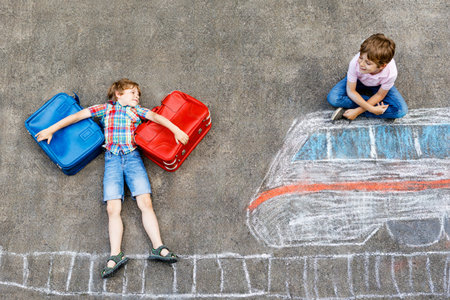 Two little kids boys having fun with picture drawing with colorful chalks on asphalt. Children having fun with chalk and crayon painting. going on vacations and travel. Friends with suitcases.の写真素材