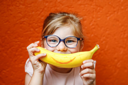 Happy little child girl with yellow banana like smile on orange background. Preschool girl with glasses smiling. Healthy fruits for children.の写真素材