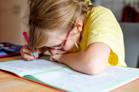 Cute elementary school girl writing in her workbook and doing school homework. Focused child learning and studying indoors. Concept of education, childhood, home learningの写真素材