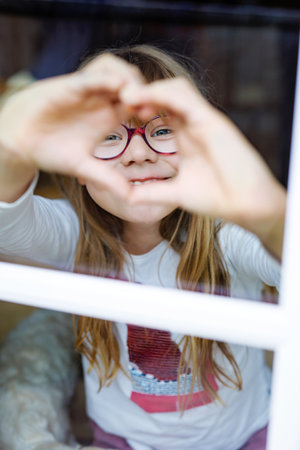 Smiling little girl with glasses making a heart shape with her hands while looking through a window, expressing love and joy in a cozy home atmosphere.の写真素材
