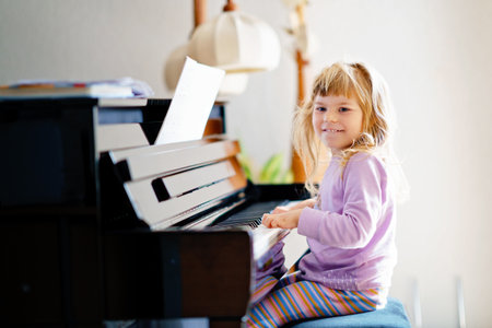 Beautiful little toddler girl playing piano in living room. Cute preschool child having fun with learning to play music instrument. Early musical education for childrenの写真素材