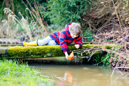 Happy little kid boy in yellow rain boots playing with paper ship boat by huge puddle on spring or autumn day. Active leisure for children. Funny child having fun outdoors, wearing colorful clothesの写真素材