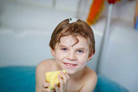 Cute little child playing with water by taking bath in bathtub at home. Adorable happy healthy preschool kid boy having fun, washing hair and head and splashing with soap.の写真素材