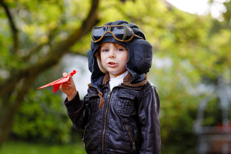 Happy little kid boy in pilot helmet and uniform playing with red toy airplane. Smiling preschool child dreaming and having fun. Education, profession, dream concept.の写真素材