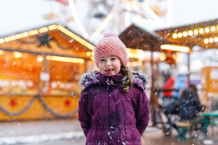 Little cute kid girl having fun on traditional Christmas market during strong snowfall. Happy child enjoying traditional family market in Germanyの写真素材