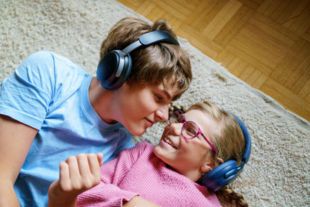 Two children, a teenage boy and a little girl, listening to music together with headphones. Smiling happy siblings enjoying sound and spending fun leisure time indoors.の写真素材
