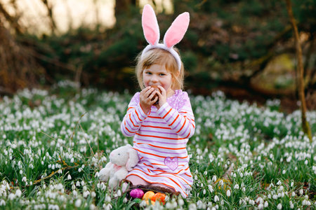 Little girl with Easter bunny ears making egg hunt in spring forest on sunny day, outdoors. Cute...