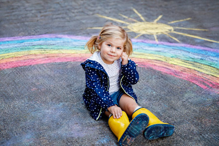 Happy little toddler girl in rubber boots with rainbow sun and clouds with rain painted with...