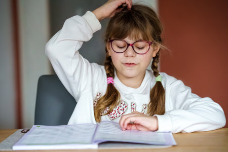 Smiling school girl with glasses writing in notebook at desk, happy child studying at home,...