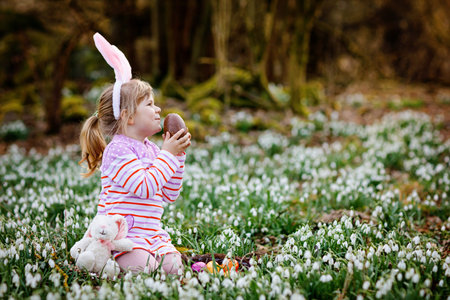 Little girl with Easter bunny ears making egg hunt in spring forest on sunny day, outdoors. Cute...