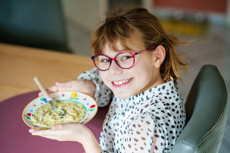 Smiling little girl with glasses eating pasta at home, cheerful child enjoying noodle soup with chicken for lunch, healthy family lifestyle conceptの写真素材