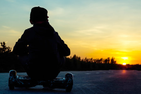 A young boy sits on a super popular electric scooter and looks at the sunset. Mini segway hoverboard...