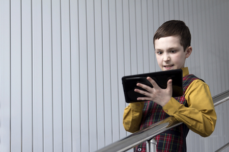 A teenager in a checkered jacket and yellow shirt looks at the tablet irritably while playing or working. White striped background.の写真素材