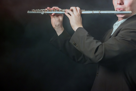 A close-up of a flute in the hands and on the lips of a musician. The guy takes the sounds on a dark background with smoke. Copy space.の写真素材