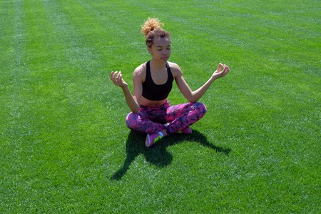 A beautiful black African-American woman in a sports black T-shirt and pink sneakers, relaxes sitting in a yoga pose and meditating on a field with green grass. Life style. Copy space.の写真素材