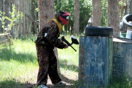 A teenager girl or boy in a spotted protective suit and wearing a red protective mask plays paintball on a forest range. Black tires, barrels with blots, like protective hiding places.の写真素材