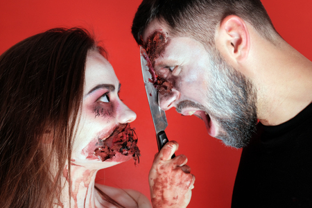 Halloween concept of makeup for All Saints or the Dead. The guy screams at the time when the girl cuts his face with a bloody knife. Horrible cosplay. Close-up. Torn wounds on the face and darned mouth.の写真素材