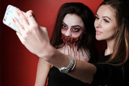 Concept selfie makeup for the story in the style of Halloween. A beautiful girl together with another girl in the image of a witch are taking pictures of themselves on the phone. Imitation of a bloody wound and a wired mouth on a woman's face. Red background.の写真素材