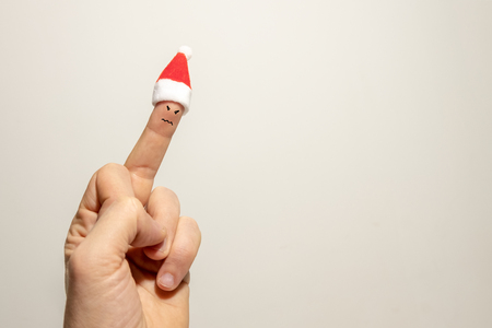 Evil emotion painted on the middle finger of a man's hand with a Santa Claus hat. The concept of a bad new year or christmas. Copy space. Gray background.の写真素材