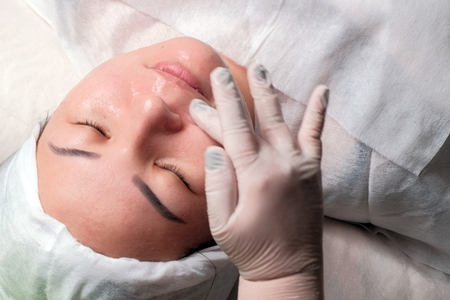 The procedure of rejuvenation and cleansing of the skin in the cosmetology center. Beautician in white gloves smears a transparent gel on the face of a girl of Asian ethnicity. Spa treatment for a young woman.の写真素材