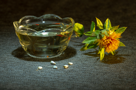 Safflower dyeing, Carthamus tinctorius, American saffron, wild thistle. Dried flower of yellow color, seeds and oil in a glass cup on a dark fabric is highlighted by a bright spot of light. Oilseed and dyeing culture since the times of ancient Egypt.の写真素材