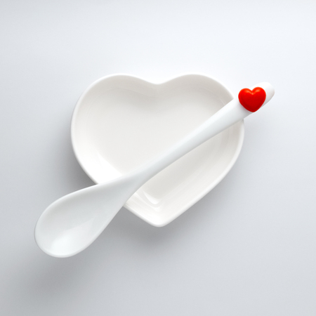 A ceramic saucer in the shape of a heart and a spoon across from it with a red small symbol of love on a white background. Glossy dishes. Object for Valentines Day or wedding romantic menu.Minimalismの写真素材