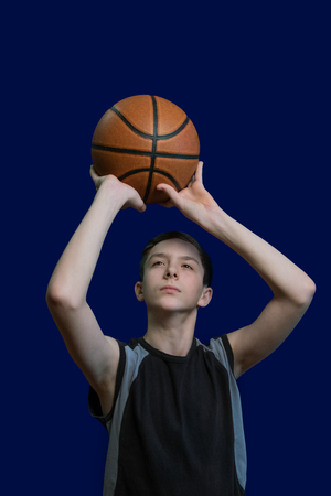 Teen basketball player. The guy in the black t-shirt holds the ball in his hands and prepares to throw it up. Template for the design of a sports poster or news about basketball competitions. Isolated on blue background.の写真素材