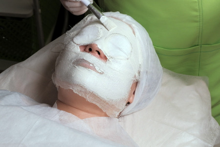 Close-up. A beautician in white gloves puts on an alginate mask with a brush on a woman's face in a beauty salon. The girl on the procedure in the cosmetology office.の写真素材